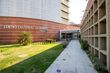 Entrada do Centro Cultural Usiminas com fachada de tijolos vermelhos e grandes janelas, um caminho pavimentado leva à porta principal.