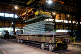 Pátio industrial da Usiminas com grandes placas de metal empilhadas sobre um vagão de transporte e sendo movidas por um guindaste.