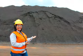 Engenheira usando capacete e colete refletivo, gesticulando em frente a uma grande pilha de minério de ferro.