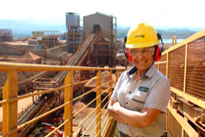 Engenheira sorridente usando capacete e óculos de segurança em uma plataforma de uma instalação industrial, com maquinário pesado ao fundo.