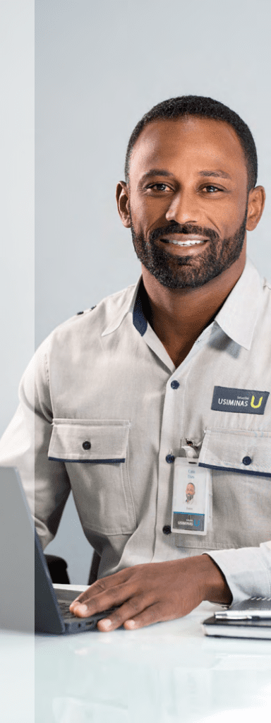 Funcionário da Usiminas, um homem negro, sorrindo para a câmera enquanto trabalha em um laptop, vestindo um uniforme de trabalho cinza com crachá visível.