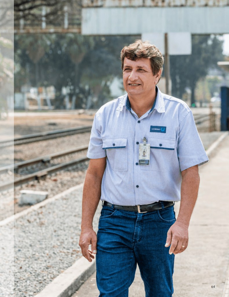 Geovani Campos Lage, técnico industrial, caminha ao longo de uma plataforma de trem, vestindo uniforme da Usiminas e sorrindo para a câmera.