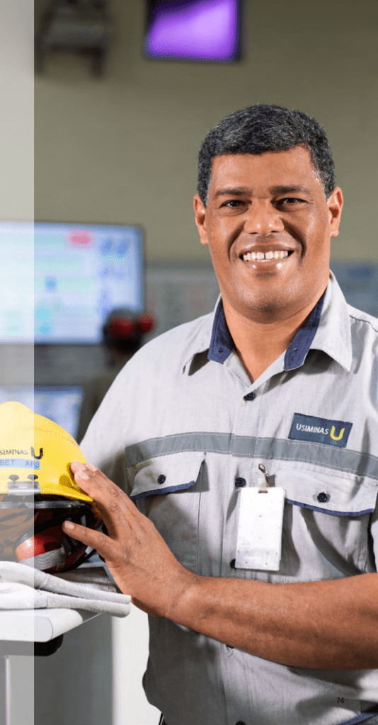 Funcionário da Usiminas, um homem negro sorridente, segurando um capacete de segurança amarelo em um ambiente de controle industrial.