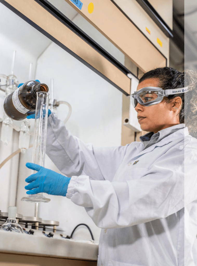 Cientista feminina trabalhando atentamente em um laboratório, usando jaleco, óculos de proteção e luvas, enquanto despeja um líquido em um recipiente de vidro.