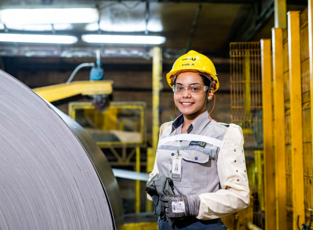 Mulher jovem com uniforme industrial, capacete amarelo e óculos de segurança, sorrindo em uma fábrica.